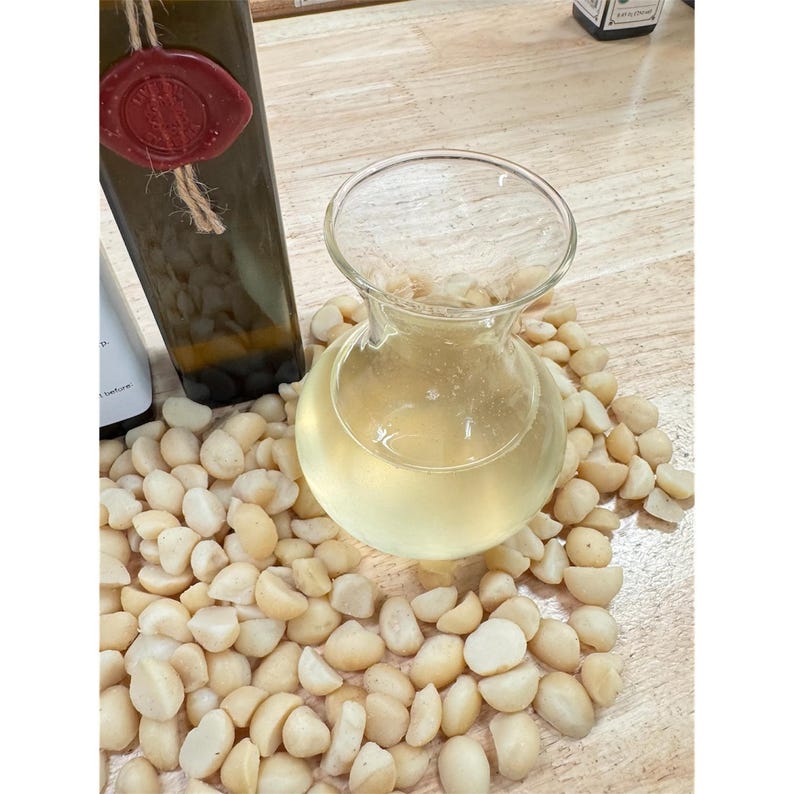 May include: A clear glass carafe filled with a light yellow liquid, surrounded by shelled macadamia nuts. A dark green bottle with a red wax seal is visible in the background. The scene is set on a light-colored wooden surface.