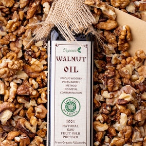 May include: A glass bottle of organic walnut oil with a burlap wrap. The label reads "Organic Walnut Oil", "Unique Wooden Press-Barrel Method", "No Metal Contamination", "Live Oil by Lesna", "100% Natural Raw First Cold Pressed", "From Organic Walnuts", "The freshest taste guaranteed by:" and "USDA Organic". The bottle is surrounded by walnut halves.
