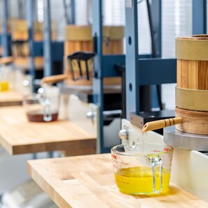 May include: A series of industrial presses featuring cylindrical wooden barrels with metal bands, mounted on blue metal frames. A clear glass measuring cup with red markings sits below a wooden spout, collecting golden liquid dripping from the press.
