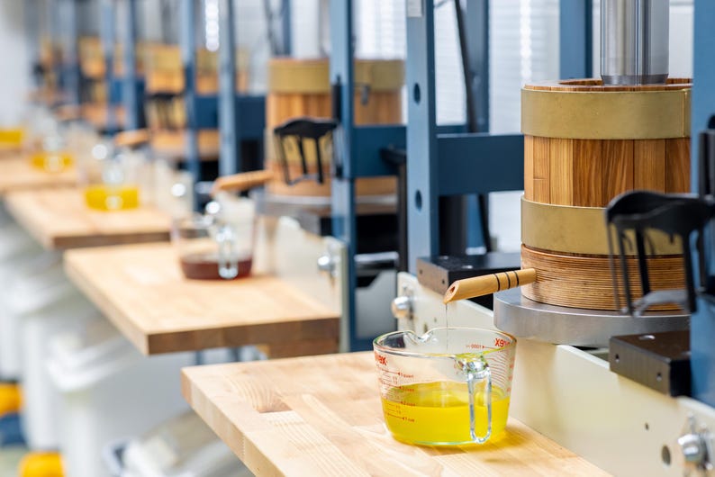 May include: A series of industrial presses featuring cylindrical wooden barrels with metal bands, mounted on blue metal frames. A clear glass measuring cup with red markings sits below a wooden spout, collecting golden liquid dripping from the press.