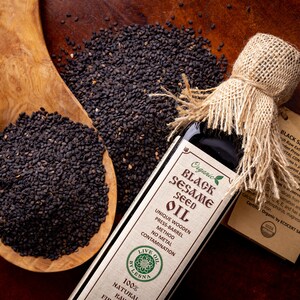 May include: A glass bottle of organic black sesame seed oil with a burlap tied around the top. The bottle is labeled with the words "Organic Black Sesame Seed Oil" and "Live Oil by Lesna". A wooden spoon filled with black sesame seeds is next to the bottle.