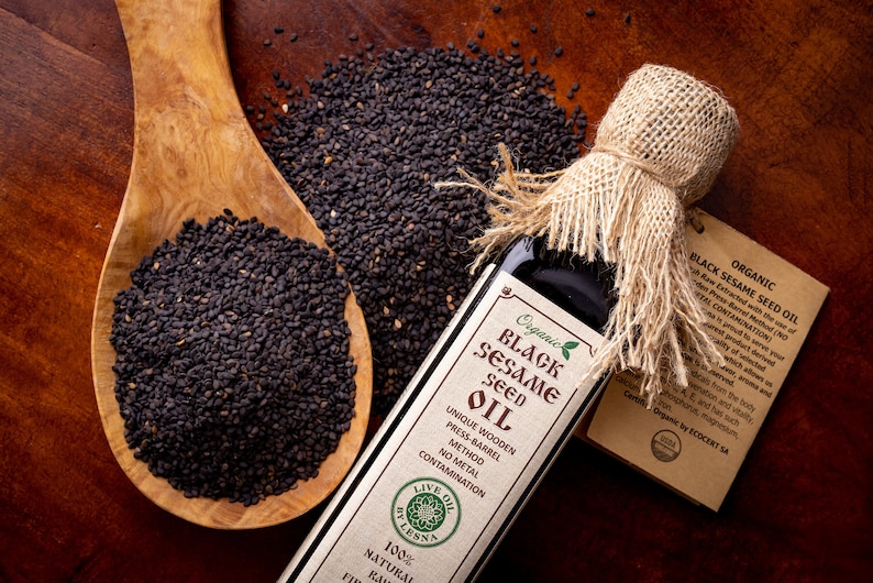May include: A glass bottle of organic black sesame seed oil with a burlap tied around the top. The bottle is labeled with the words "Organic Black Sesame Seed Oil" and "Live Oil by Lesna". A wooden spoon filled with black sesame seeds is next to the bottle.