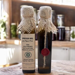 May include: Two glass bottles of pecan oil, each with a burlap-covered top and a label. The labels read "Pecan Oil from Organic Pecans" and "Raw Cold Pressed 100% Natural Premium Quality." One bottle has a red wax seal. Each bottle contains 8.45 oz.
