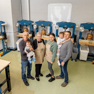 May include: A family of five stand in front of a row of industrial-style presses, likely used for making oil or juice. The adults are wearing aprons, and the youngest child is being held by one of the adults. The room is clean and well-lit, with a light green floor.