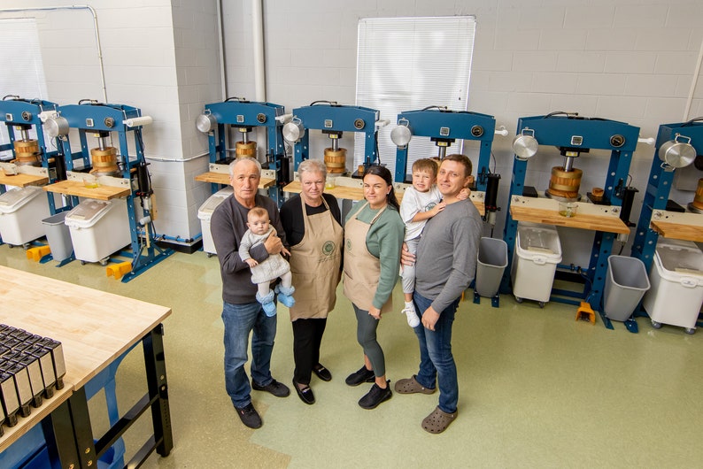 May include: A family of five stand in front of a row of industrial-style presses, likely used for making oil or juice. The adults are wearing aprons, and the youngest child is being held by one of the adults. The room is clean and well-lit, with a light green floor.
