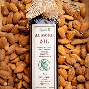 May include: A glass bottle of organic almond oil with a hessian tied around the top. The bottle is labelled "Organic Almond Oil" and features a green leaf logo. The bottle is surrounded by almonds. The label also states "Unique Wooden Press-Barrel Method, No Metal Contamination, Live Oil by Lesna, 100% Natural Raw First Cold Pressed, From Organic Almonds, The freshest taste guaranteed by: USDA Organic, 8.45 oz (250 ml)"