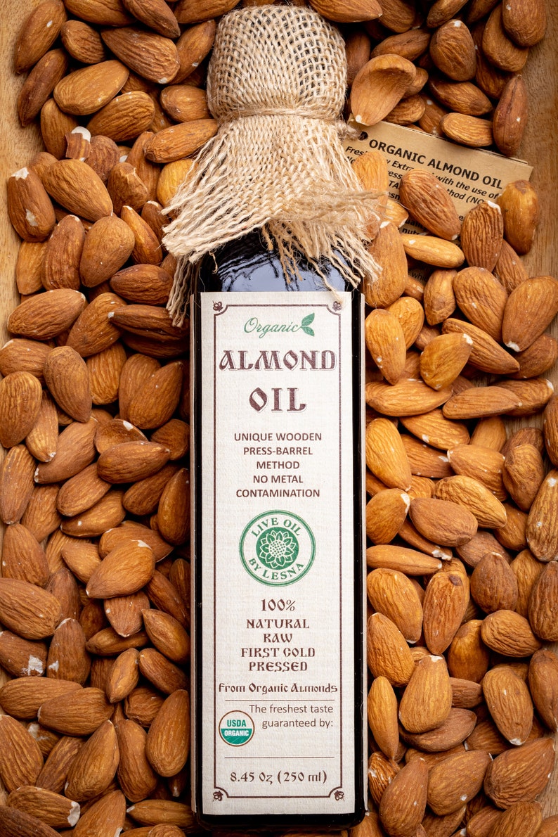 May include: A glass bottle of organic almond oil with a hessian tied around the top. The bottle is labelled "Organic Almond Oil" and features a green leaf logo. The bottle is surrounded by almonds. The label also states "Unique Wooden Press-Barrel Method, No Metal Contamination, Live Oil by Lesna, 100% Natural Raw First Cold Pressed, From Organic Almonds, The freshest taste guaranteed by: USDA Organic, 8.45 oz (250 ml)"