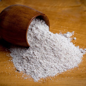 May include: A dark brown wooden bowl tipped on its side, spilling a mound of fine, light beige powdery substance onto a matching wooden surface. The texture of the powder appears granular and dry, contrasting with the smooth, polished wood grain.