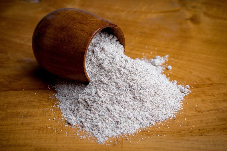 May include: A dark brown wooden bowl tipped on its side, spilling a mound of fine, light beige powdery substance onto a matching wooden surface. The texture of the powder appears granular and dry, contrasting with the smooth, polished wood grain.