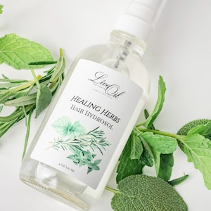 May include: A clear glass bottle with a white spray top and a white label. The label says "Loved Oil by Lorna Family Oil Mill" and "Healing Herbs Hair Hydrosol". The bottle is surrounded by green leaves.