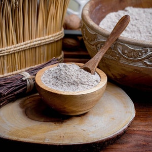 May include: A small wooden bowl filled with light brown flour and a wooden spoon, resting on a circular wooden slice. Nearby are a bundle of thin dark twigs, a larger carved wooden bowl containing more flour, and a tall woven straw container.