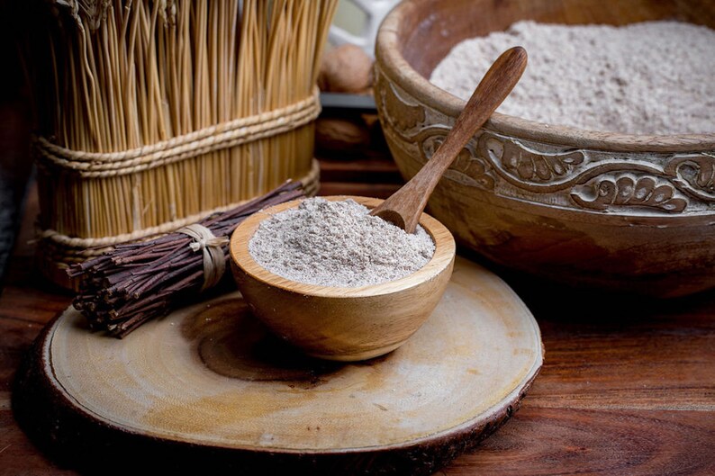 May include: A small wooden bowl filled with light brown flour and a wooden spoon, resting on a circular wooden slice. Nearby are a bundle of thin dark twigs, a larger carved wooden bowl containing more flour, and a tall woven straw container.