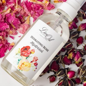 May include: A clear glass bottle with a white spray top and a white label. The label has a floral design and the text "Live Oil by Leana Family Oil Well Hydrating Rose Hydrosol". The bottle is surrounded by dried rose petals.
