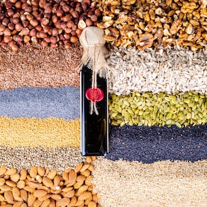 May include: A variety of nuts and seeds arranged in rows, including hazelnuts, flax seeds, poppy seeds, mustard seeds, chia seeds, almonds, sunflower seeds, pumpkin seeds, walnuts, and a bottle of dark oil with a red wax seal.