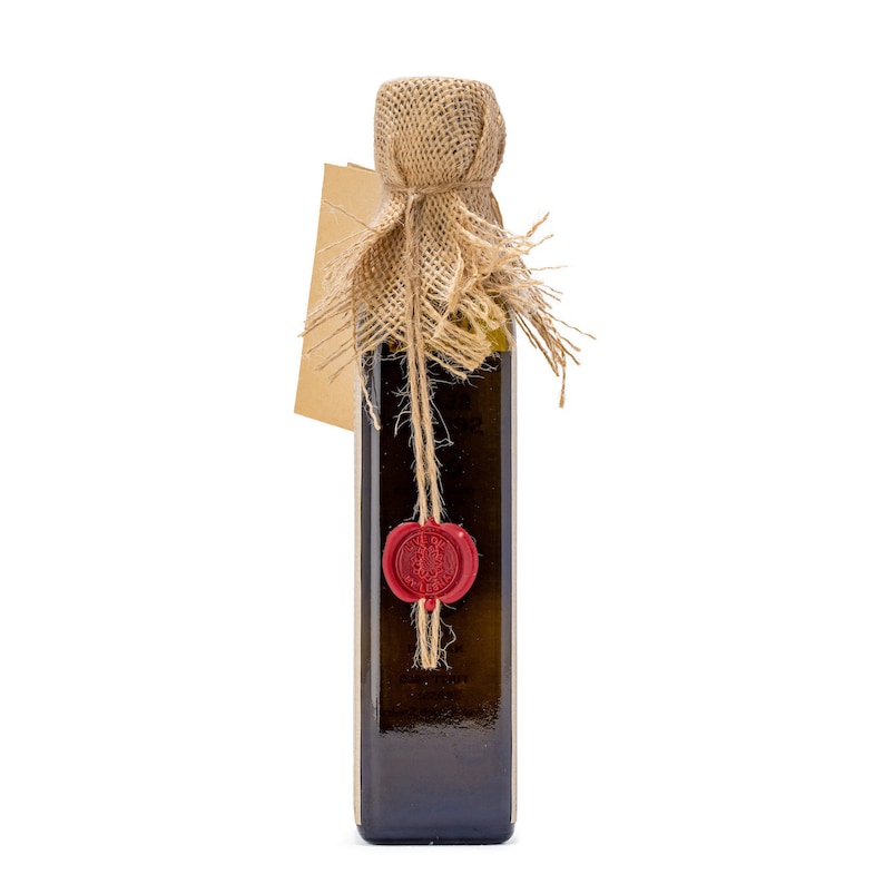 May include: A glass bottle of olive oil with a burlap sack covering the top of the bottle. The bottle has a red wax seal with a design on it. The bottle is sitting on a white background.