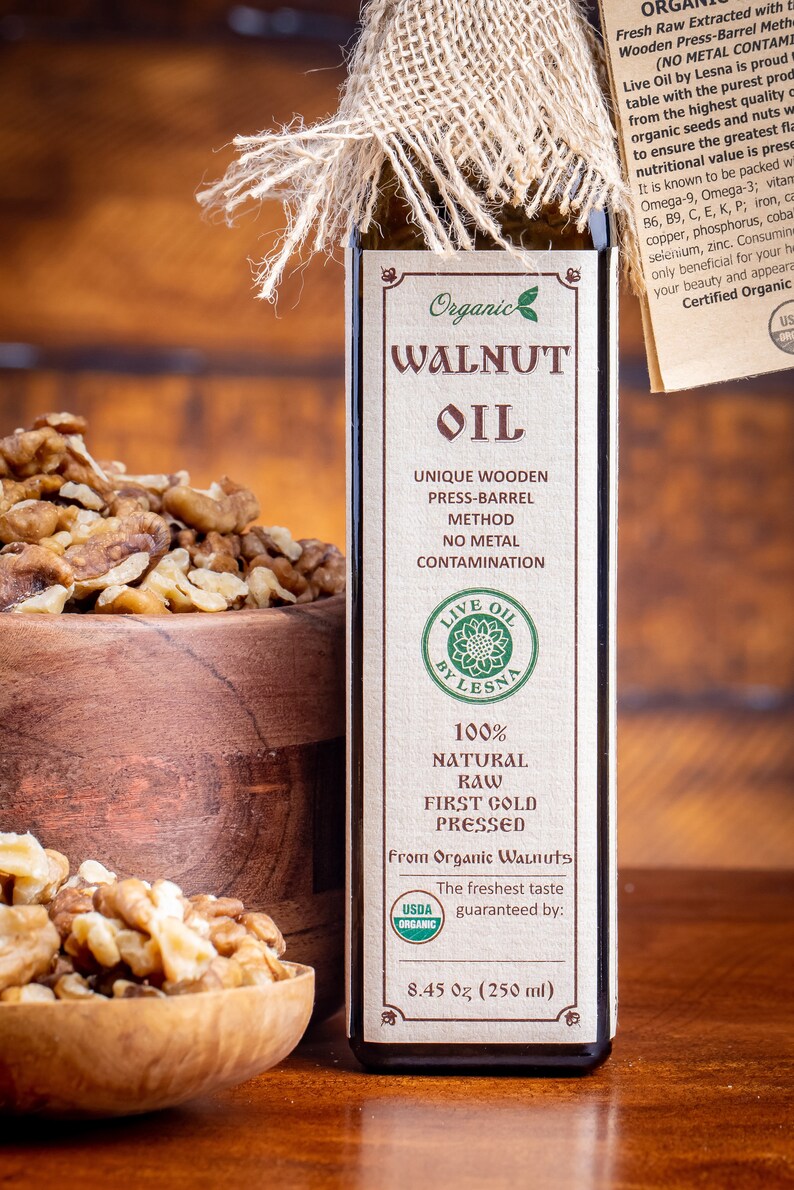 May include: A glass bottle of organic walnut oil with a brown label. The label says "Organic Walnut Oil", "Unique Wooden Press-Barrel Method", "No Metal Contamination", "Live Oil by Lesna", "100% Natural Raw First Cold Pressed", "From Organic Walnuts", "The freshest taste guaranteed by:", and "8.45 oz (250 ml)". There is a USDA Organic seal on the label. The bottle is sitting on a wooden surface next to a bowl of walnuts.