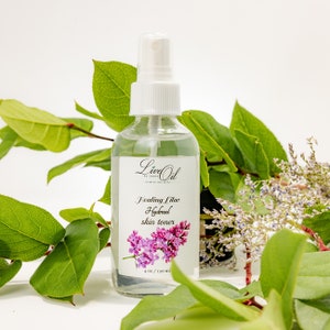 May include: A clear glass spray bottle of Healing Lilac Hydrosol skin toner, 4 oz / 120 ml, with a white cap and label featuring a lilac flower illustration. The bottle is surrounded by green leaves and white flowers.