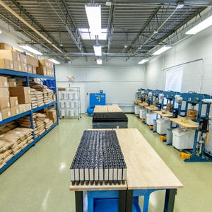 May include: A manufacturing facility interior featuring tall blue metal shelving units stocked with white boxes and brown paper bags. A central wooden workbench holds rows of black rectangular components, while blue industrial presses line the wall.