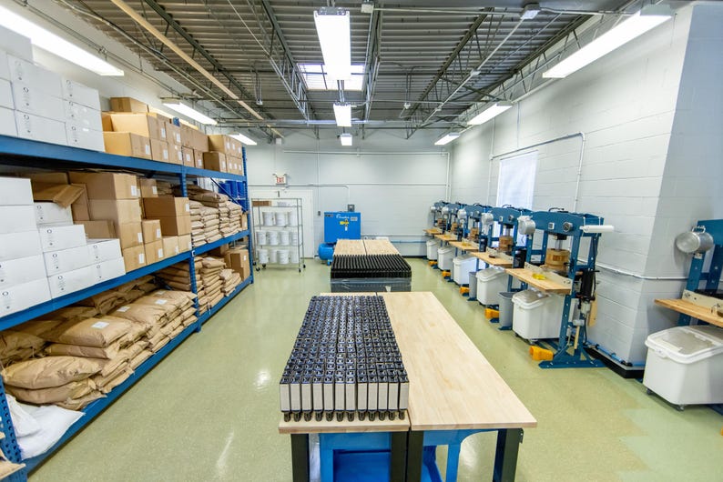 May include: A manufacturing facility interior featuring tall blue metal shelving units stocked with white boxes and brown paper bags. A central wooden workbench holds rows of black rectangular components, while blue industrial presses line the wall.