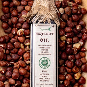 May include: A rectangular glass bottle of hazelnut oil with a burlap-wrapped cap, surrounded by raw hazelnuts. The label reads Organic Hazelnut Oil, Live Oil By Lesna, 100% Natural Raw First Cold Pressed, From Organic Hazelnuts, 8.45 oz (250 ml).
