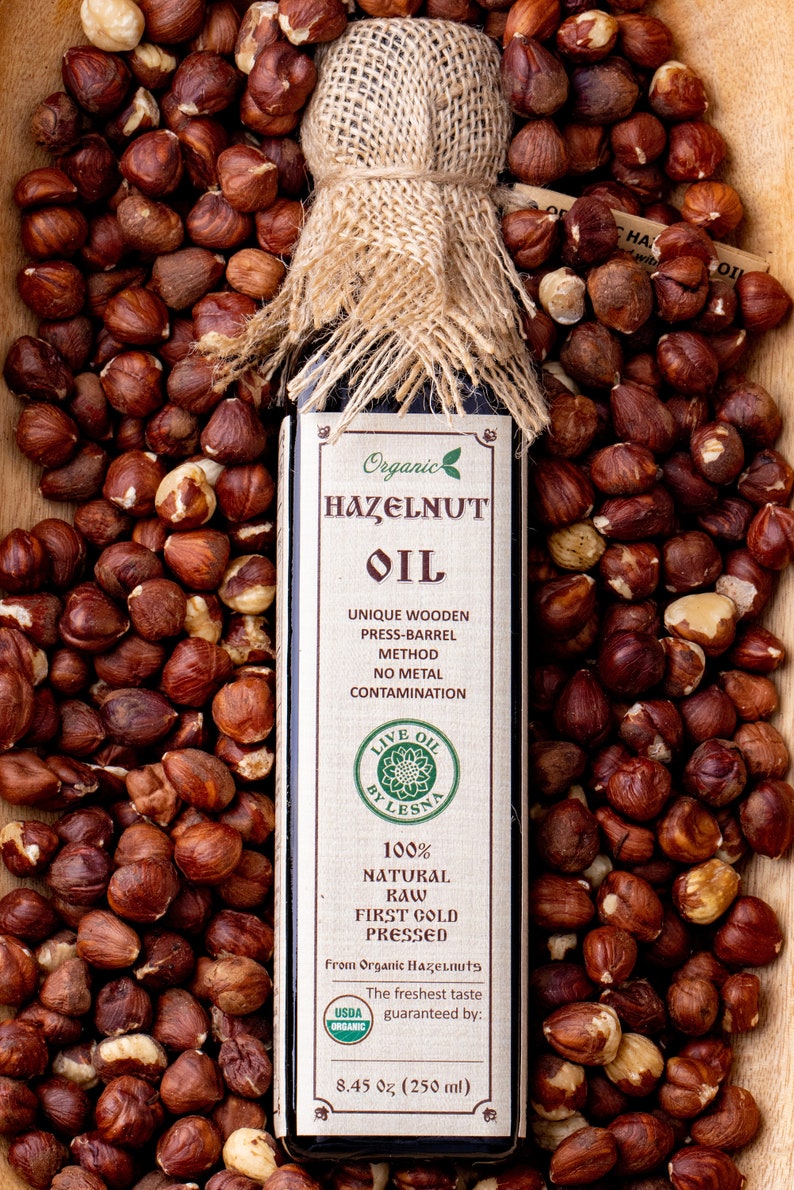 May include: A rectangular glass bottle of hazelnut oil with a burlap-wrapped cap, surrounded by raw hazelnuts. The label reads Organic Hazelnut Oil, Live Oil By Lesna, 100% Natural Raw First Cold Pressed, From Organic Hazelnuts, 8.45 oz (250 ml).