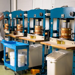 May include: An industrial setting with multiple blue and gold machinery. A person in a lab coat and hair net stands near a blue cart. Each machine has a wooden component and a small glass container with a yellow liquid.