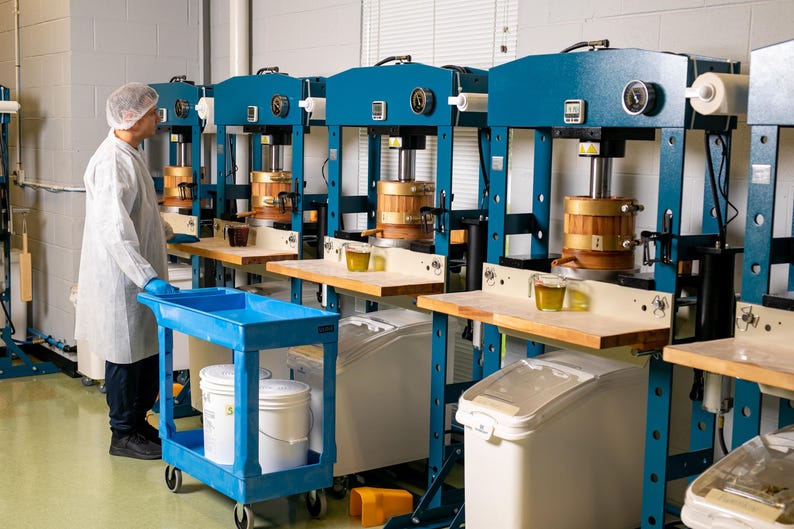 May include: An industrial setting with multiple blue and gold machinery. A person in a lab coat and hair net stands near a blue cart. Each machine has a wooden component and a small glass container with a yellow liquid.