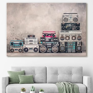 May include: A collection of vintage boomboxes stacked on top of each other against a textured gray wall. The boomboxes are in various colors and styles, including black, silver, and red. Some of the boomboxes have cassette tape players, while others have CD players.