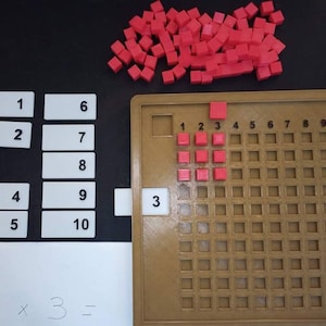 May include: A brown multiplication grid with red blocks showing the equation 3 x 3 = 9. The grid is numbered 1 through 10. There are also separate number cards from 1 to 10.