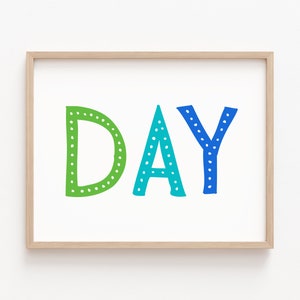 May include: A framed print with the word "DAY" in large, colorful letters. The letters are green, teal, and blue, with white dot details. The frame is a light wood color, and the background is white.