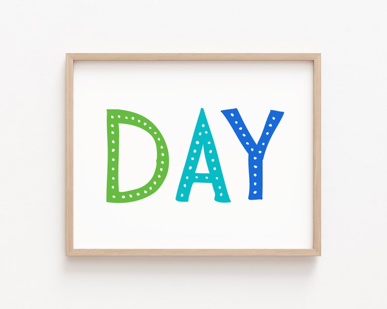 May include: A framed print with the word "DAY" in large, colorful letters. The letters are green, teal, and blue, with white dot details. The frame is a light wood color, and the background is white.