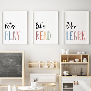 May include: Three white framed prints with the text "let's" in black and the words "PLAY", "READ", and "LEARN" in rainbow colors. The prints are hanging on a white wall above a play kitchen and a chalkboard.