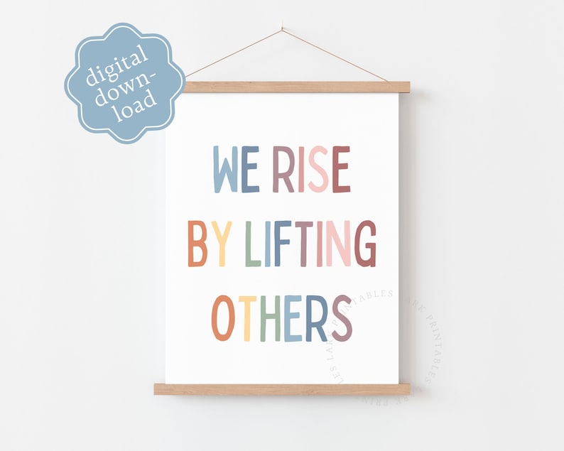 May include: A white poster with the text "WE RISE BY LIFTING OTHERS" in colorful block letters. The poster is held by wooden bars at the top and bottom and is hanging from a string. A blue digital download badge is in the upper left corner.