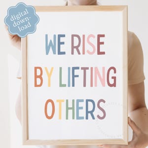 May include: A framed print with the words "WE RISE BY LIFTING OTHERS" in colorful letters. The print has a light wood frame and a light blue digital download sticker in the upper left corner. The text is in various colors.