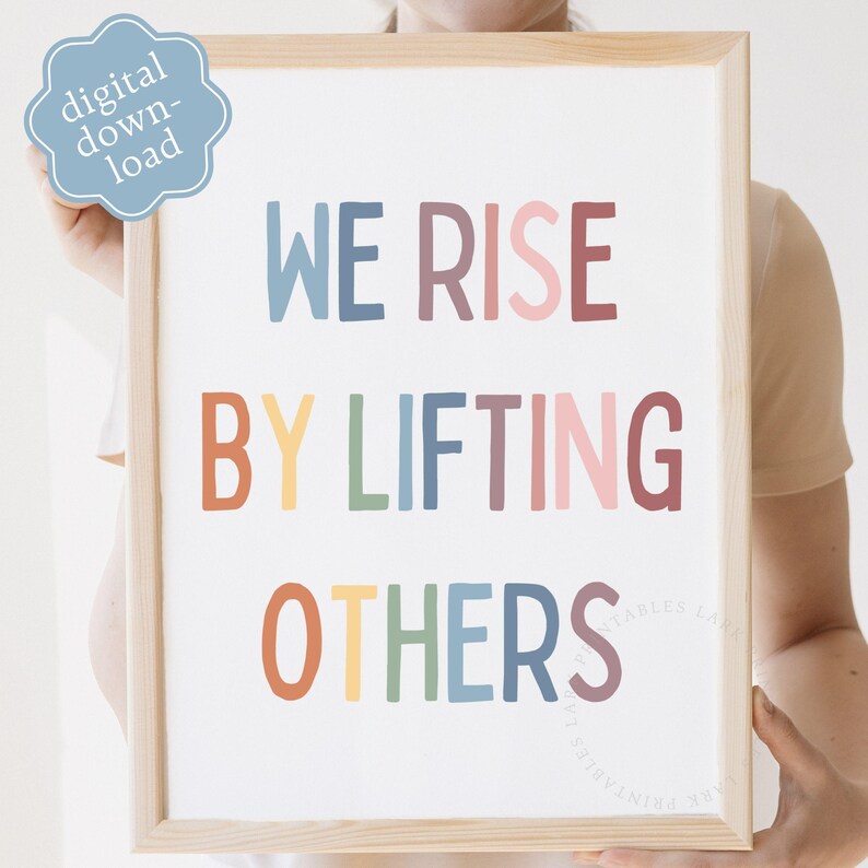 May include: A framed print with the words "WE RISE BY LIFTING OTHERS" in colorful letters. The print has a light wood frame and a light blue digital download sticker in the upper left corner. The text is in various colors.