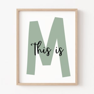 May include: A framed print featuring a large, sage green letter M and the text "This is" in black cursive font.
