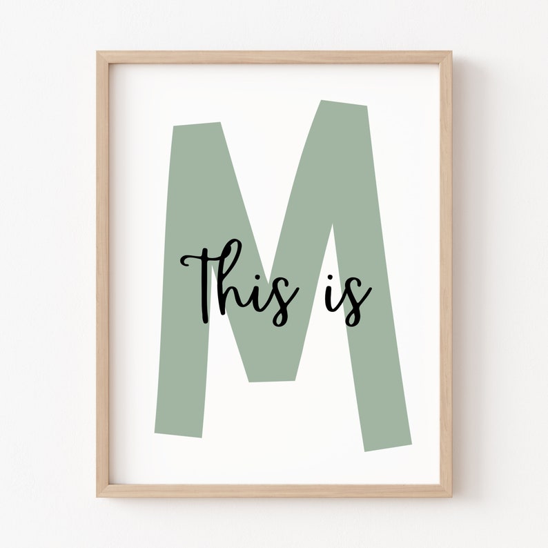 May include: A framed print featuring a large, sage green letter M and the text "This is" in black cursive font.