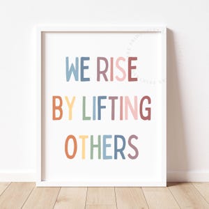 May include: A framed print with the words "WE RISE BY LIFTING OTHERS" in colorful, playful lettering. The text is in blue, red, yellow, green, pink, orange, and purple. The print is in a white frame.