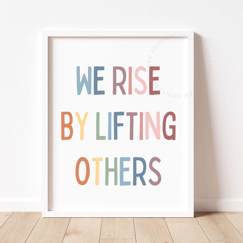May include: A framed print with the words "WE RISE BY LIFTING OTHERS" in colorful, playful lettering. The text is in blue, red, yellow, green, pink, orange, and purple. The print is in a white frame.
