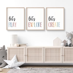May include: Three framed prints with the text "let's play", "let's explore", and "let's create" in colorful letters. The prints are hung on a white wall above a light wood cabinet with three doors.
