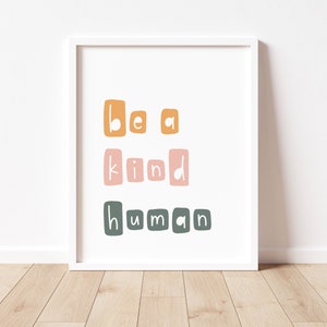 Puede incluir: Una impresi&oacute;n art&iacute;stica enmarcada con la frase "be a kind human" en una fuente l&uacute;dica y en bloques. El texto est&aacute; dispuesto en tres l&iacute;neas, con cada palabra en un color diferente: naranja, rosa y verde. La impresi&oacute;n est&aacute; sobre un fondo blanco.