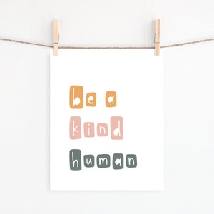Puede incluir: Una impresi&oacute;n blanca con las palabras "be a kind human" en letras coloridas y cuadradas. La impresi&oacute;n est&aacute; colgada de un tendedero con pinzas de madera. Las letras son en tonos de naranja, rosa y verde.