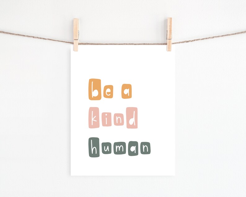 Puede incluir: Una impresi&oacute;n blanca con las palabras "be a kind human" en letras coloridas y cuadradas. La impresi&oacute;n est&aacute; colgada de un tendedero con pinzas de madera. Las letras son en tonos de naranja, rosa y verde.