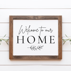 Puede incluir: Un letrero de madera enmarcado con las palabras "Welcome to our HOME" en letras negras. Una ramita decorativa de hojas está debajo de la palabra HOME. El letrero está sobre un fondo blanco con textura de vetas de madera.