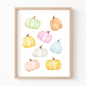 May include: A framed print featuring nine watercolor pumpkins in various pastel colors, including light green, orange, pink, yellow, white, and blue. The pumpkins are arranged in a random pattern on a white background.