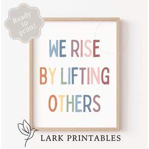 May include: A framed print with the text "WE RISE BY LIFTING OTHERS" in colorful, playful lettering. The print is in a light wood frame and includes the text "Ready to print!" and "LARK PRINTABLES".