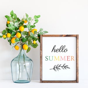 May include: A wooden frame with a white background and the text "hello SUMMER" in colorful letters. The frame is sitting on a white surface next to a clear glass vase filled with a green leafy plant and yellow lemons.