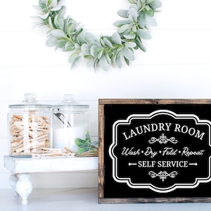 Pode incluir: Placa preta enquadrada com texto branco que diz "Laundry Room, Wash, Dry, Fold, Repeat, Self Service". A placa está sobre um fundo de madeira branca.