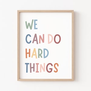 May include: A framed print with the text "WE CAN DO HARD THINGS" in colourful, playful lettering. The background is white, and the frame is a light wood colour.