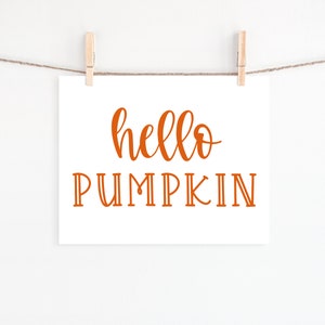 May include: A white card with the text "hello pumpkin" written in orange cursive lettering. The card is hanging from a string with two wooden clothespins.